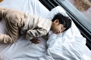 Een jong kind in pyjama slaapt op een groot wit bed bij het raam