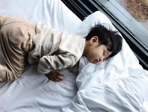 Een jong kind in pyjama slaapt op een groot wit bed bij het raam