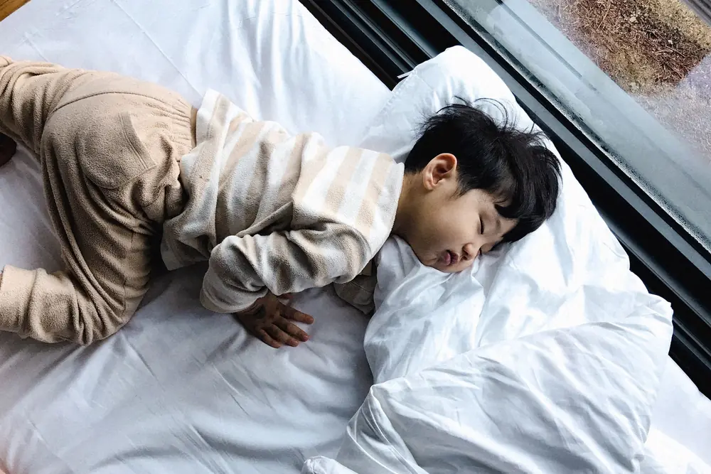 Een jong kind in pyjama slaapt op een groot wit bed bij het raam