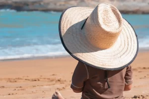 Een peuter met een grote strohoed zit op het strand en kijkt naar de zee